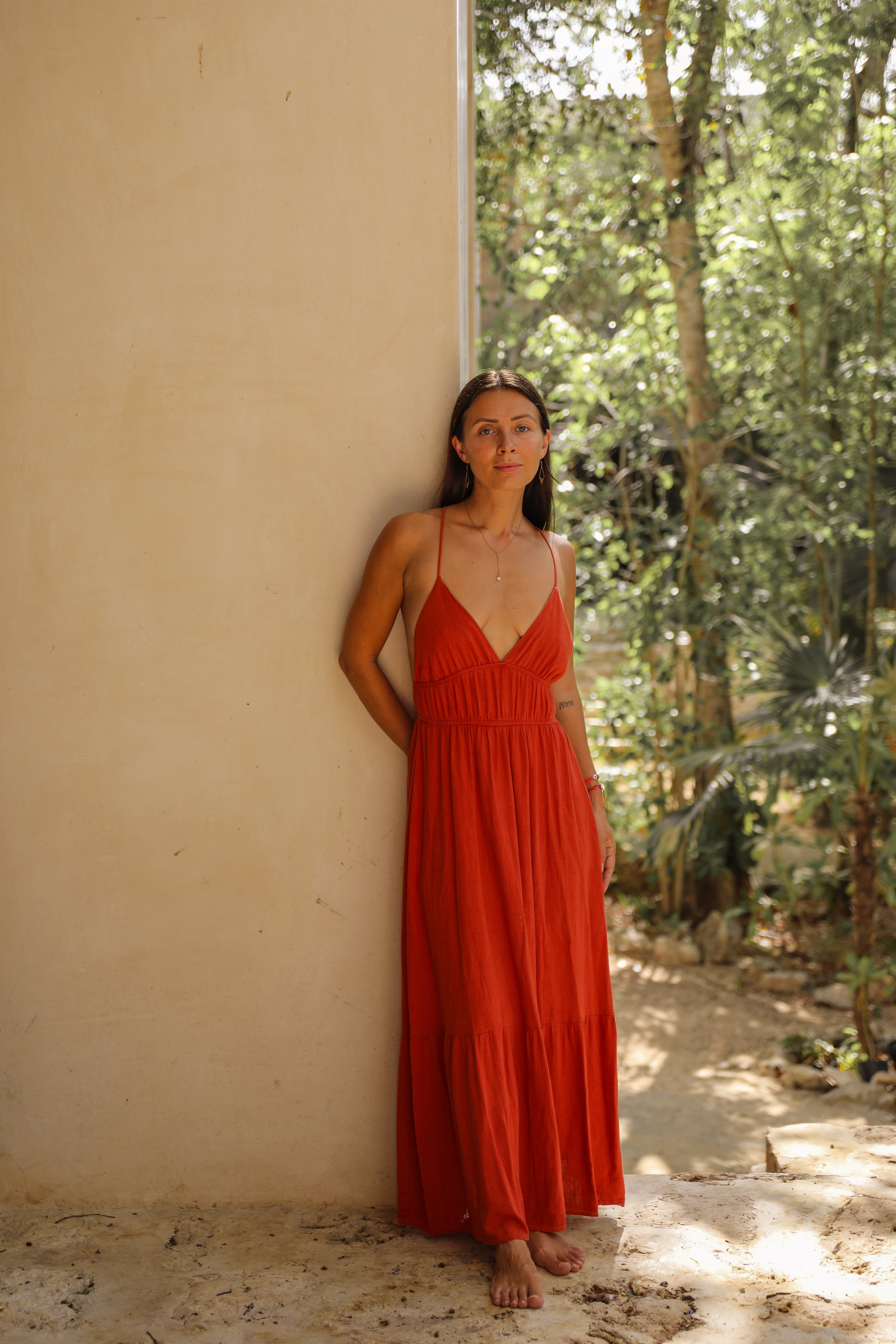 Zelata in a red dress, leaning on a wall in a garden